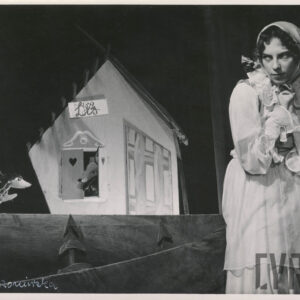 z okna małego domku spogląda kukiełka przedstawiająca lisa, obok stoi jeż, po prawej stronie kobieta w długiej, białej sukni z chustą na głowie, ręce trzyma blisko ciała, złożone jak do modlitwy