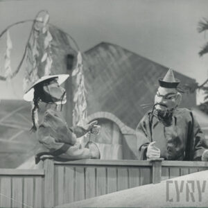 drewniany płotek, na nim siedzi lalka w chińskim kapeluszu z długimi wąsami i warkoczem, obok stoi druga lalka w stożkowej czapeczce z wąsami i brodą, za nimi budynek i drzewa