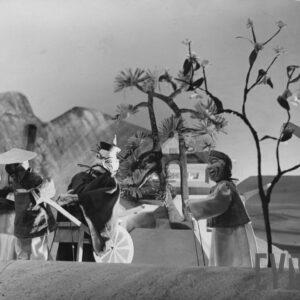 z lewej strony lalka w chińskiej czapce wiezie w pieszej rikszy lalkę z wąsami i brodą, obok stoi lalka w długiej szacie, za nimi drzewa, fragment domu i góry