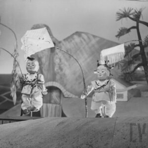 trzy lalki dzieci stoją obok siebie trzymając w rękach latawce, za nimi drzewa i góra