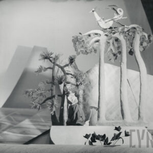 pośrodku sceny stoi lalka z siekierą w ręku, obok znajduje się drzewo, na którym siedzi okazała kukiełka przedstawiająca ptaka, z tyłu inne drzewa i góra