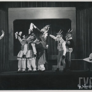 aktorzy w maskach kozłów machają białymi chustami do stojącego naprzeciw innego aktora w masce kozła, siedzący po prawej stronie aktor ma na kolanach księgę a w ręce pióro  