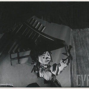 zza scenografii wychyla się lalka ubrana w koszulę i spódnicę, lewą ręką opiera się o element dekoracji