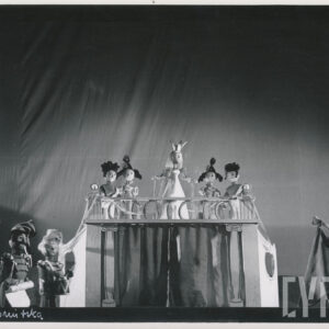 pięć lalek na podwyższeniu, stojąca pośrodku ma na głowie koronę i ubrana jest w długą suknię, poniżej lalka przedstawiająca koziołka ubrana w długą pelerynę, po drugiej stronie trzy lalki w mundurach z instrumentami