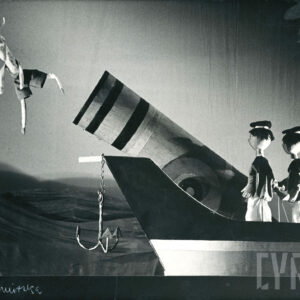 scenografia przedstawia statek z działem, u którego wylotu znajduje się lalka przedstawiająca koziołka, na pokładzie stoją odwrócone tyłem dwie lalki w strojach marynarzy