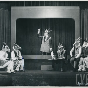 pośrodku sceny aktor w masce kozła z uniesioną prawą ręką, po obu stronach siedzą na krzesłach aktorzy w maskach kozłów, jeden z nich ma na kolanach księgę i w ręce trzyma pióro, po lewej stronie aktor w masce kozła z aparatem fotograficznym