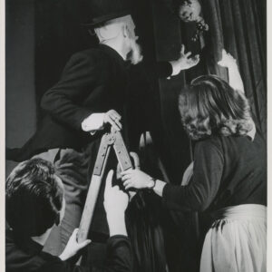 na szczeblach drabiny, po obu jej stronach stoją kobieta i mężczyzna wyciągający ręce w kierunku kotary, zza której wychyla się lalka, obok drabiny stoi drugi mężczyzna 