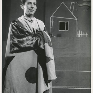 przed czterostopniowymi schodami stoi mężczyzna okryty długą peleryną