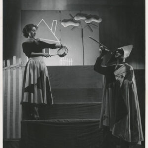 na pierwszym stopniu schodów stoi kobieta w długiej spódnicy z apaszką na szyi, w rękach trzyma bębenek i pałeczkę, przy schodach stoi mężczyzna w długiej pelerynie i nakryciu głowy, prawą ręką przy ustach trzyma trąbkę 