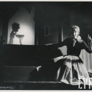 na stopniu schodów siedzi kobieta w długiej spódnicy i apaszce na szyi, unosi w górę lewą rękę z wyprostowanym palcem wskazującym, za nią scenografia przedstawiająca dom z lalką wewnątrz