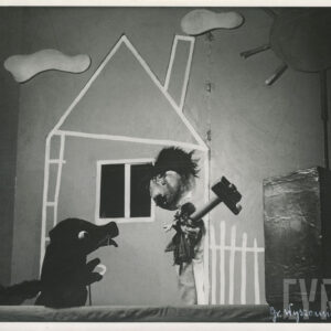 na scenie znajdują się dwie lalki, z których jedna przedstawia postać psa, a druga mężczyznę, który w rękach trzyma młotek, za nimi elementy scenografii, dom, chmury i słońce