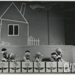 na scenie znajduje się sześć lalek w różnych pozach, za nimi elementy scenografii, dom, chmury i słońce