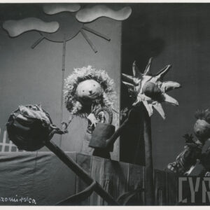 na scenie znajdują się dwie lalki, jedna przedstawia dziewczynę z długimi warkoczami, która trzyma w rękach konewkę, druga przedstawia chłopca, za nimi elementy scenografii, fragment domu, chmury i słońce