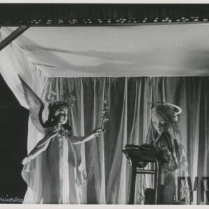 lalka w bardzo długich, rozpuszczonych włosach i aureoli stoi przy pulpicie, na którym leży książka, obok lalka w długiej sukni ze skrzydłami na plecach oraz kwiatami we włosach i lewej dłoni, z tyłu dekoracje