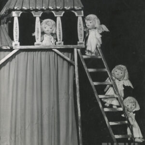 trzy lalki ubrane w długie suknie ze skrzydłami na plecach, stoją na szczeblach drabiny, czwarta lalka stoi na balkonie pod daszkiem 