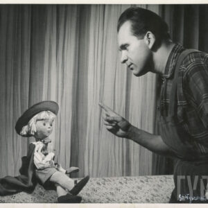 mężczyzna ubrany w spodnie na szelkach i koszulę w kratę, wskazuje palcem prawej ręki na siedzącą obok lalkę, ubraną w pelerynę i kapelusz