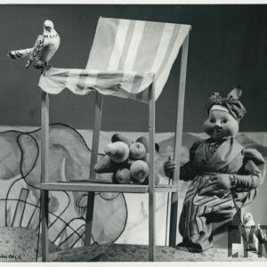 przy straganie, na którym leżą owoce, siedzi na ławeczce lalka ubrana w długa suknię i fartuch, na głowie nosi chustę, obok niej i na dachu straganu znajdują się kukiełki przedstawiające gołębie