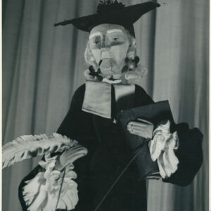 lalka ubrana w togę i biret, w lewej ręce trzyma księgę, a w prawej białe pióro