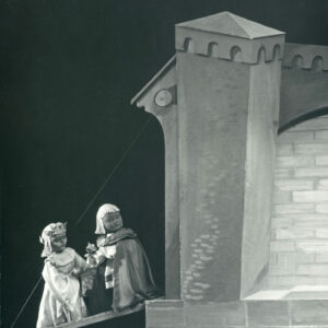 przed zamkiem na moście zwodzonym stoją dwie lalki, jedna ubrana w długą suknię, druga ma na sobie pelerynę