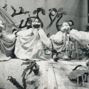 scena w sadzie, cztery lalki w długich sukniach i chustach na głowach stoją obok siebie z rozpostartymi ramionami, z przodu rosną pojedyncze warzywa, z tyłu jabłonie i fragment budynku