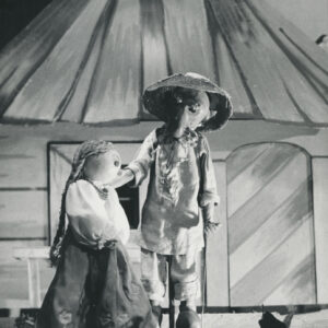 scena przed domem, na scenie dwie lalki, starszy mężczyzna w kapeluszu i chuście na szyi rozmawia z dziewczyną z długim warkoczem ubraną w białą koszulę, długą spódnicę i korale