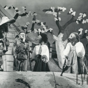 w sadzie za domem stoją obok siebie cztery lalki - starszy mężczyzna w kapeluszu, młody elegancko ubrany chłopak oraz dwie dziewczyny ubrane w białe bluzki i długie spódnice, za nimi kwitnąca jabłoń, drewniany płot i fragment budynku