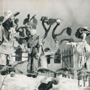 scena w sadzie, stoją dwie lalki - starszy mężczyzna w kapeluszu i drugi z wąsami, w koszuli w kratę i młotkiem w ręku, za nimi strach na wróble, jabłoń i drewniany płot