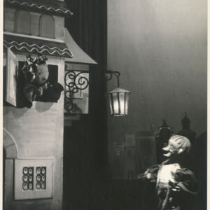 z okna kamienicy wychyla się lalka z glinianym dzbankiem w ręce, przed budynkiem stoi druga lalka w okularach i długim płaszczu