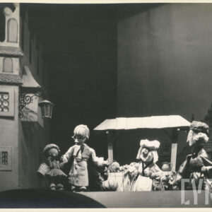 scena przed kamienicą, przy straganie z warzywami stoją cztery lalki w różnych pozach