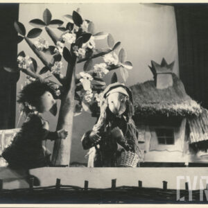 przed domem pokrytym strzechą znajdują się dwie lalki, jedna trzyma w rękach koszyk i laskę, za nimi kwitnące drzewo