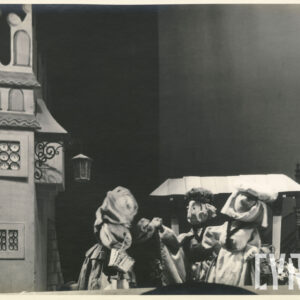 scena przed kamienicą, przy straganie stoi lalka w długiej białej sukni i nakryciu głowy, obok dwie inne lalki wyciągają ku sobie ręce