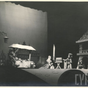 na scenie przed kamienicą znajdują się cztery lalki, jedna z nich chowa się za straganem, z prawej strony stoi wieża 