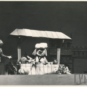 za straganem z warzywami stoi lalka w nakryciu głowy, obok stoi druga lalka z wyciągniętą przed siebie ręką