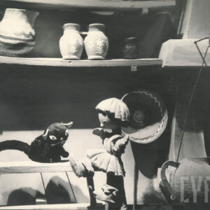 scena wewnątrz domu, na ławce siedzi czarny kot, obok niego lalka przedstawiająca chłopca, nad nimi półka z glinianymi dzbankami