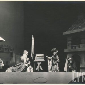 na scenie obok wieży stoją dwie elegancko ubrane lalki, pomiędzy nimi znajduje się stół, przy wieży pochylona lalka w długiej pelerynie