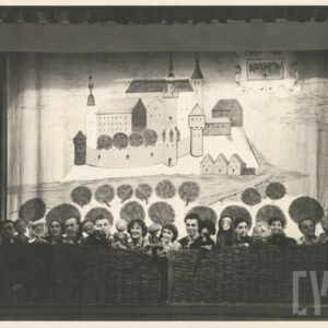 zza dekoracji spoglądają aktorzy trzymający w rękach kukiełki, za nimi rozwieszona tkanina z malowidłem zabudowań Krakowa