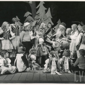 na scenie siedzi mężczyzna z długą brodą, ubrany w płaszcz i czapkę, na lewym kolanie trzyma worek z prezentami, wokół niego siedzą i stoją dzieci w strojach regionalnych