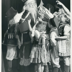 mężczyzna z długą broda ubrany w płaszcz i czapkę stoi otoczony dziećmi, które podnoszą ręce w górę  