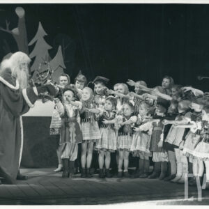 po lewej stronie sceny stoi mężczyzna z brodą ubrany w długi płaszcz, obok stoi duża grupa dzieci w strojach regionalnych, które wyciągają ręce w jego stronę 