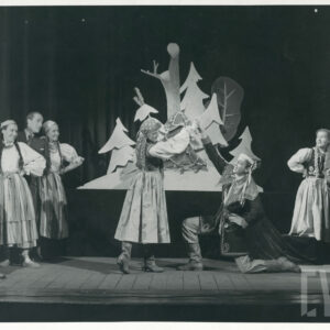 na scenie mężczyzna przyklęka na lewym kolanie, prawą rękę unosi wysoko chwytając dłoń kobiety w długiej sukni, po obu stronach pary stoją i siedzą kobiety oraz mężczyźni w regionalnych strojach