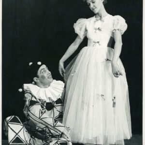 na scenie stoi kobieta trzymając w dłoniach materiał swojej długiej sukni z kokardkami, obok siedzi mężczyzna z pomalowaną twarzą w stroju pajaca 