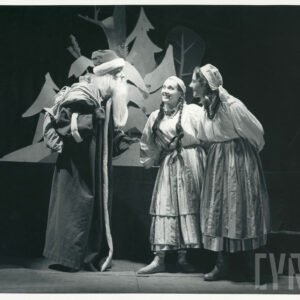 na scenie stoi mężczyzna z siwą brodą i workiem na plecach, ubrany w długi płaszcz, obok stoją dwie kobiety w białych bluzkach i długich spódnicach 