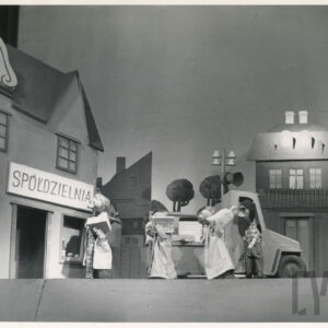 scena w miasteczku, przed budynkiem z napisem spółdzielnia stoi auto dostawcze i lalka w czapce i koszuli w kratę, obok stoją jedna za drugą trzy lalki z pakunkami w rękach, zwrócone w kierunku sklepu  