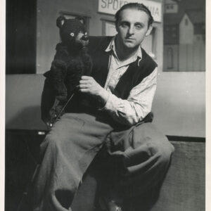 na scenie przysiadł aktor ubrany w szerokie spodnie, koszulę w kratę i kamizelkę, w rękach trzyma lalkę misia