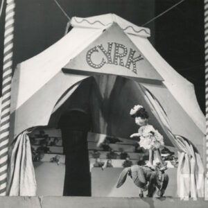 lalka ubrana w sukienkę z falbankami i kokardą na głowie stoi na kukiełce przedstawiającej osiołka, z tyłu namiot cyrkowy z widownią i kukiełkami