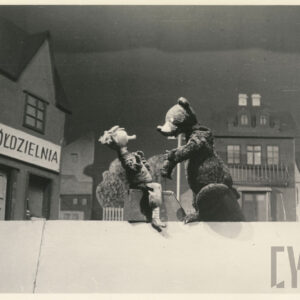 scena w miasteczku, przed budynkiem z napisem spółdzielnia siedzą naprzeciw siebie dwie lalki - miś z wyciągniętymi przed siebie łapami i mężczyzna, który się od niego odchyla