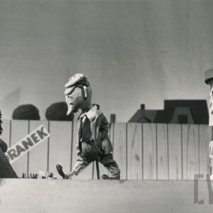 na podnóżku, przed którym leży kilka okrągłych pudełek w paski, siedzi lalka trzymająca w lewej ręce szczotkę , obok stoi lalka mężczyzny w eleganckim ubraniu z krawatem, za nimi drewniany płot i słup ogłoszeniowy oklejony plakatami  