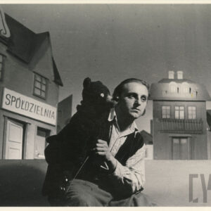 na scenie siedzi aktor ubrany w koszulę w kratę i kamizelkę, w rękach trzyma kukiełkę przedstawiającą niedźwiedzia, za nim scenografia z różnymi budynkami