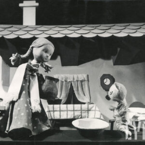 scenografia przedstawia wnętrze pokoju, na scenie znajdują się dwie lalki - dziewczynka w długiej sukience w kropki trzymająca dzbanek i kot, przed którym stoi miska