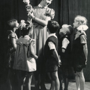 na scenie stoi aktorka trzymająca lalkę przedstawiającą kota, wokół stoi pięcioro dzieci ze zwróconymi w jej kierunku twarzami